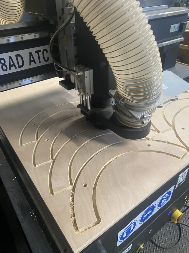 CNC router cutting out next day complex semicircular shapes in 18mm plywood, with two holes in each